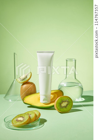 A cosmetic tube without label standing on a half of avocado, displayed with some glassware and a petri dish of kiwi slices. Natural beauty blank label for branding mock-up concept 114767357