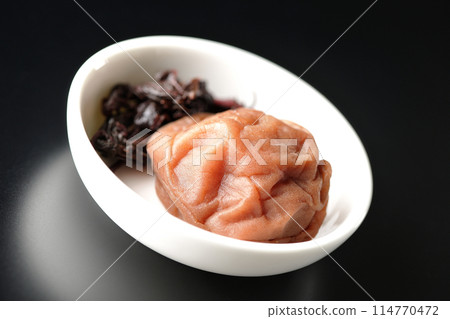 Red shiso and pickled plums served on a small plate photographed against a black background 114770472