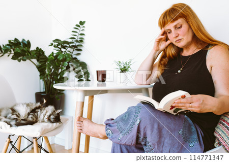 portrait middle-aged blonde woman sitting at table with book in home interior 114771347