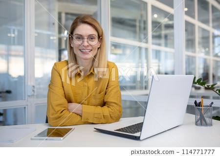 Smiling woman in a modern office setting, sitting at a desk with a laptop, wearing glasses and a yellow blouse with confidence. 114771777