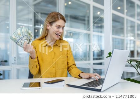 Woman in a yellow blouse holding cash and using a laptop in a bright, modern office, expressing excitement and success. 114771803