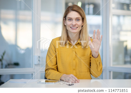 Smiling woman in a yellow shirt waves hello during a video call in an office environment, expressing friendliness and warmth. 114771851