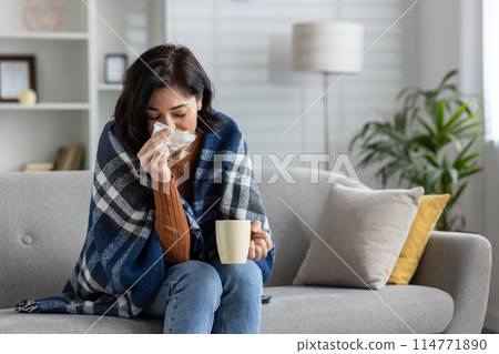 Young woman sitting at home on the sofa and sick, covered with a blanket, wiping her nose from a cold with a napkin, holding a cup with a drink and suffering from a virus. Young woman sitting at home on the sofa and sick, covered with a blanket, wiping her nose from a cold with a napkin, holding a cup with a drink and suffering from a virus. 114771890
