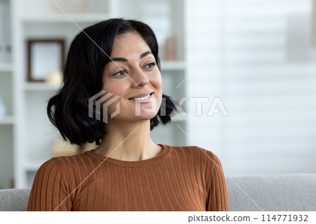 Smiling woman sitting on sofa in living room, looking thoughtful and content. Cozy home environment 114771932