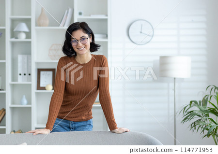Smiling woman wearing glasses and casual clothes standing confidently in a bright home office environment. 114771953