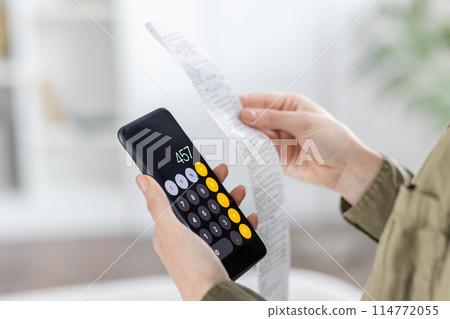 A midsection view of a woman using a calculator and examining a long receipt in a bright, modern room. She manages household finances, ensuring budget accuracy. A midsection view of a woman using a calculator and examining a long receipt in a bright, modern room. She manages household finances, ensuring budget accuracy. 114772055