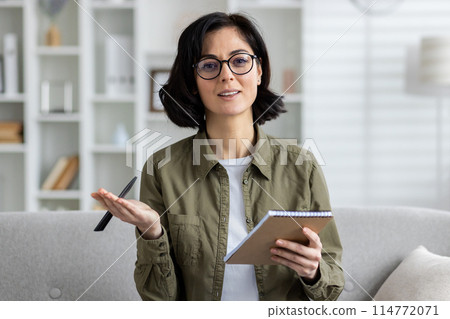 A confident woman wearing glasses holds a pen and notebook while discussing and looking at the camera, likely engaged in an online chat or virtual class session from her bright living room. 114772071