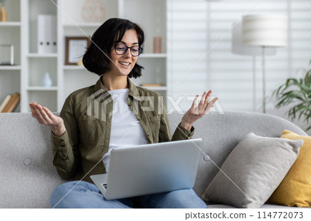 A young smiling woman in casual clothes is sitting on the sofa at home, holding a laptop on her lap and talking on a video call while gesturing with her hands. A young smiling woman in casual clothes is sitting on the sofa at home, holding a laptop on her lap and talking on a video call while gesturing with her hands. 114772073