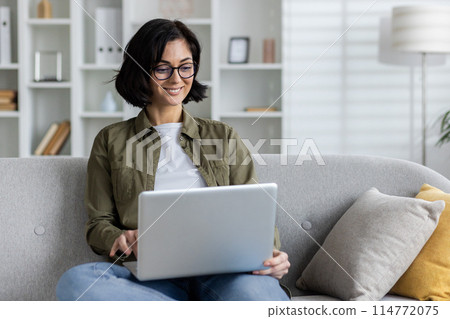 A cheerful adult woman in glasses works on her laptop from a comfortable sofa in a well-lit modern living room, exhibiting productivity and relaxation at home. A cheerful adult woman in glasses works on her laptop from a comfortable sofa in a well-lit modern living room, exhibiting productivity and relaxation at home. 114772075