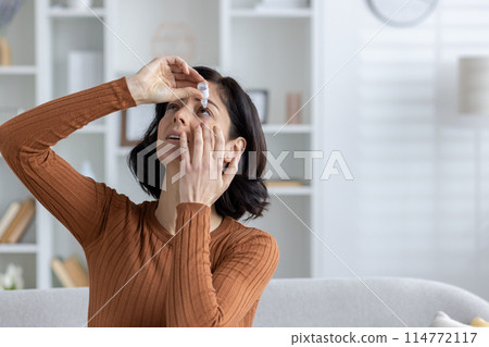 Close-up photo of a young woman sitting on the sofa at home and instilling medicine for fatigue and inflammation in her eyes. 114772117