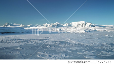 Fly over snowy mountain landscape in Antarctica. Polar frozen ocean landscape covered by snow under blue sunny sky. Discover the beauty of South Pole. Antarctica travel and exploration background 114775742