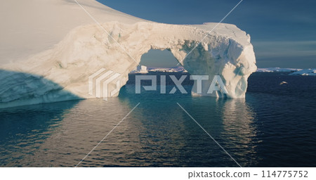 Melted ice cave arch hole iceberg towering arctic ocean. Big glacier floating cold water in sunset. Ecology, melting ice, climate change and global warming concept. Drone flight close up Melted ice cave arch hole iceberg towering arctic ocean. Big glacier floating cold water in sunset. Ecology, melting ice, climate change and global warming concept. Drone flight close up 114775752
