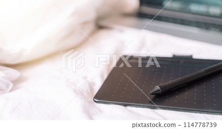Graphic tablet with a pen in front of a laptop on a white bed 114778739