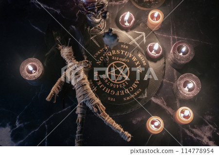 A haunting occult setup featuring a pendulum, mystical symbols, candles, a voodoo doll, and a ritual goblet amidst swirling smoke. 114778954