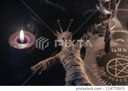 A haunting occult setup featuring a pendulum, mystical symbols, candles, a voodoo doll, and a ritual goblet amidst swirling smoke. 114778955