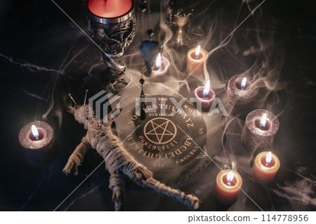 A haunting occult setup featuring a pendulum, mystical symbols, candles, a voodoo doll, and a ritual goblet amidst swirling smoke. 114778956