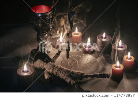 A haunting occult setup featuring a pendulum, mystical symbols, candles, a voodoo doll, and a ritual goblet amidst swirling smoke. 114778957