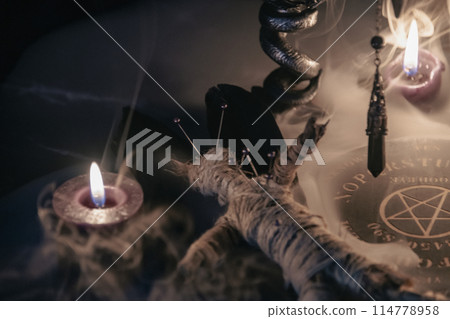 A haunting occult setup featuring a pendulum, mystical symbols, candles, a voodoo doll, and a ritual goblet amidst swirling smoke. A haunting occult setup featuring a pendulum, mystical symbols, candles, a voodoo doll, and a ritual goblet amidst swirling smoke. 114778958