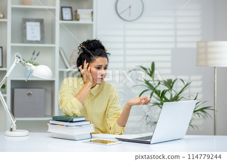 Woman feeling frustrated while working on a laptop in a home office setting with books and paperwork. Woman feeling frustrated while working on a laptop in a home office setting with books and paperwork. 114779244