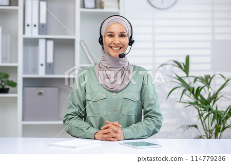 A cheerful woman in a hijab wearing a headset, working in a modern office, demonstrates professionalism and customer support. A cheerful woman in a hijab wearing a headset, working in a modern office, demonstrates professionalism and customer support. 114779286