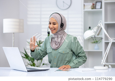 Smiling woman wearing a hijab using a laptop for a video call while working from home in a modern office setup. Smiling woman wearing a hijab using a laptop for a video call while working from home in a modern office setup. 114779290