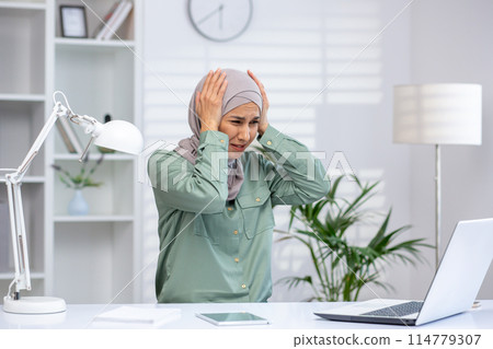 A stressed businesswoman wearing a hijab working at her office desk with a laptop, displaying an overwhelmed expression. Concept of stress, frustration, and work pressure. 114779307