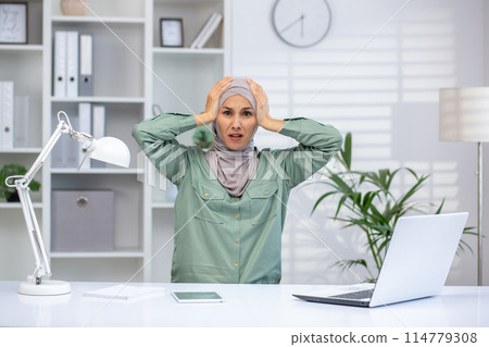 Stressed woman in hijab holding head in frustration while working in modern office. Concept of workplace anxiety, stress, and dealing with professional challenges. 114779308