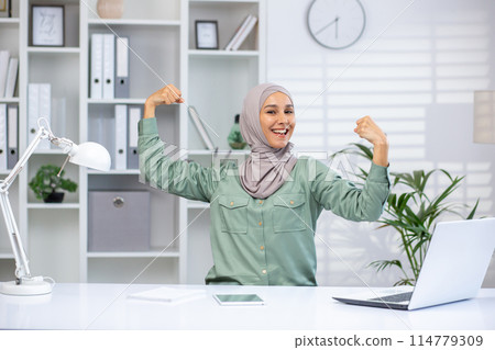 Confident businesswoman in hijab flexing muscles and smiling while working at desk in modern office environment. 114779309