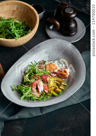 Salad with crab and avocado on a dark background. Arugula salad leaves seasoned with sauce on white wine with crab meat and grilled avocado in soft focus. Top view 114780010