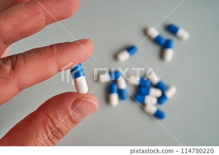 A hand holds a white and blue pill on a blue background. 114780220