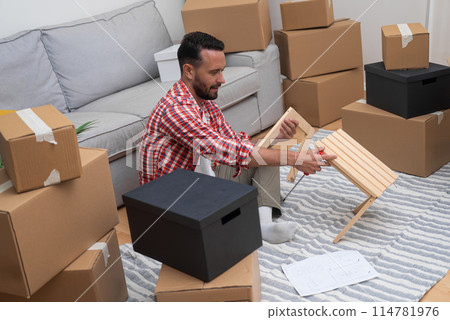 Smilling man sits with wooden furniture among various carton boxes with stuff for interior decoration enjoying moving in new contemporary house in another town Smilling man sits with wooden furniture among various carton boxes with stuff for interior decoration enjoying moving in new contemporary house in another town 114781976