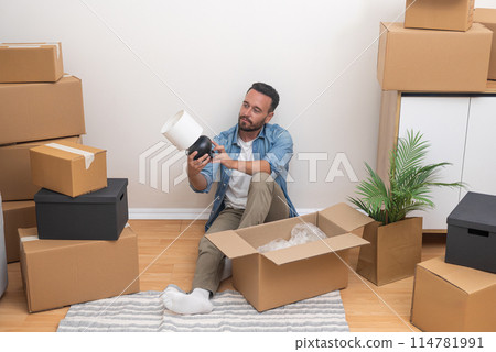 In the midst of relocation, a young man unboxing a lamp from among unpacked boxes in his new apartment, determined to make it home 114781991