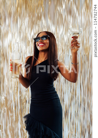 Young happy fashion African girl black woman in cocktail dress and sunglasses holding drink glass and wine bottle dancing at party with glitter background having fun at holiday, birthday celebration. 114782744