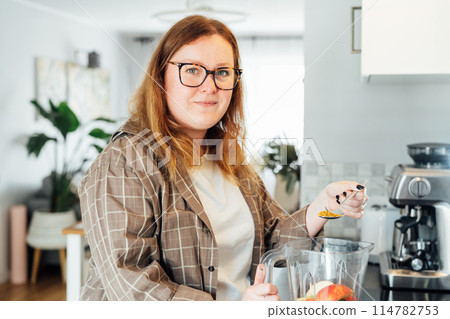 Plus size young woman adding bee pollen during making smoothie on the kitchen. Superfood supplement. Healthy detox vegan diet. Healthy dieting eating, weight loss program. Selective focus 114782753