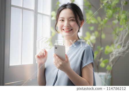 一位女士在窗邊拿著智慧型手機,看著鏡頭思考 一位女士在窗邊拿著智慧型手機,看著鏡頭思考 114786212