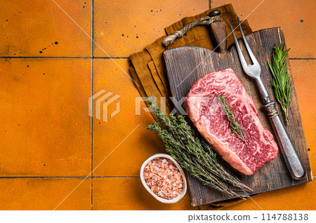 Raw Wagyu striploin, club steak on wooden board with thyme and rosemary ready for cooking. Orange background. Top view. Copy space Raw Wagyu striploin, club steak on wooden board with thyme and rosemary ready for cooking. Orange background. Top view. Copy space 114788138