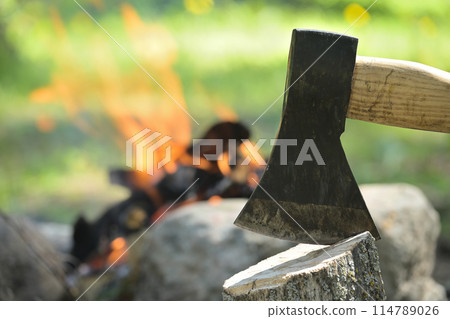 Hatchet embedded in a log of wood opposite campfire 114789026