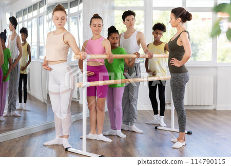 Teenage girl mastering first ballet position at barre with group Teenage girl mastering first ballet position at barre with group 114790115