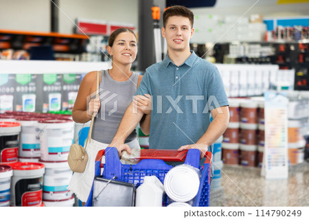 Smiling young woman and man with trolley looking for paints for house renovation and talking friendly while visiting construction hypermarket Smiling young woman and man with trolley looking for paints for house renovation and talking friendly while visiting construction hypermarket 114790249