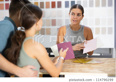 Female saleswoman of paintwork material shop advises spouses buyer with samples and product catalog 114790299