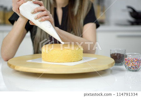 Closeup hand piping white cream on a sponge cake, creating a decorative pattern on the top of a round, light-colored cake that's placed on a wooden turntable or cake stand. Closeup hand piping white cream on a sponge cake, creating a decorative pattern on the top of a round, light-colored cake that's placed on a wooden turntable or cake stand. 114791334