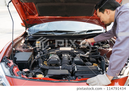 A mechanic in uniform and protective glove looks at a sport car's engine to fix it. Working atmosphere at car repair shop. A mechanic in uniform and protective glove looks at a sport car's engine to fix it. Working atmosphere at car repair shop. 114791434