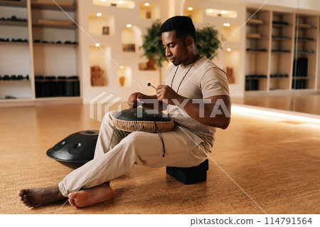 Happy calm black drummer male in trance enjoying playing using drumsticks on glucophone sitting cross-legged on floor in cozy meditation room. Concept of mindfulness, tranquility, harmony. Happy calm black drummer male in trance enjoying playing using drumsticks on glucophone sitting cross-legged on floor in cozy meditation room. Concept of mindfulness, tranquility, harmony. 114791564