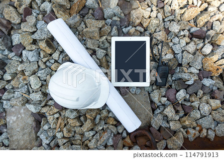 A tablet computer, hardhat, walkie-talkie and train route chart lie on ballast stone in railroad next to railroad sleeper and rail beam. Top view 114791913