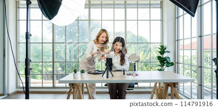 Two asian women creating content for social media. They are using a studio setup that include a smartphone mounted on a tripod, lighting equipment, and various makeup products arrayed on the table. Two asian women creating content for social media. They are using a studio setup that include a smartphone mounted on a tripod, lighting equipment, and various makeup products arrayed on the table. 114791916