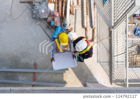 Two worker in hardhat and reflective vest are busy, looking at blueprint on a sunny day at a construction site. High angle view. 114792117