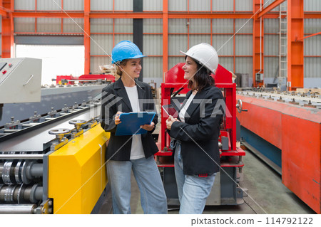 Two women with safety helmet and black suit standing in a factory, holding a clipboard and tablet computer, used for taking note, checking schedule, reviewing digital document, or communication. Two women with safety helmet and black suit standing in a factory, holding a clipboard and tablet computer, used for taking note, checking schedule, reviewing digital document, or communication. 114792122