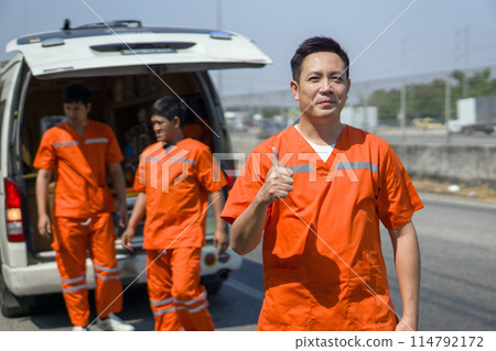 Asian rescue staff in orange uniform stand in front of an ambulance with open door and colleague working behind. Urgent assistance during road accident. 114792172