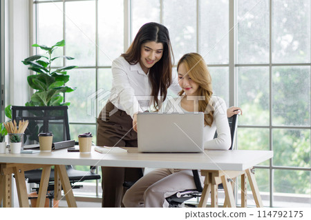 Two asian women working on laptop computer with a coffee cup, mobile phone and document folder at a desk. The background is a glass wall, overlooking the scenery of green nature 114792175