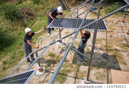Three men solar installers wearing safety helmets and work overalls while mounting photovoltaic solar panel system on sunny day. Male workers installing solar module on metal construction. 114793011